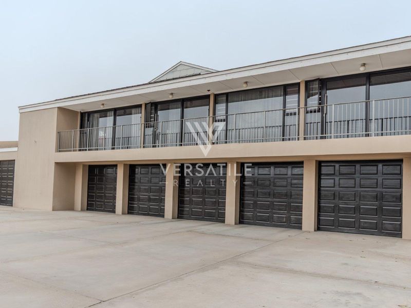 A modern exterior view featuring a spacious concrete driveway and a five-car garage with an upper-level balcony and sliding glass doors.