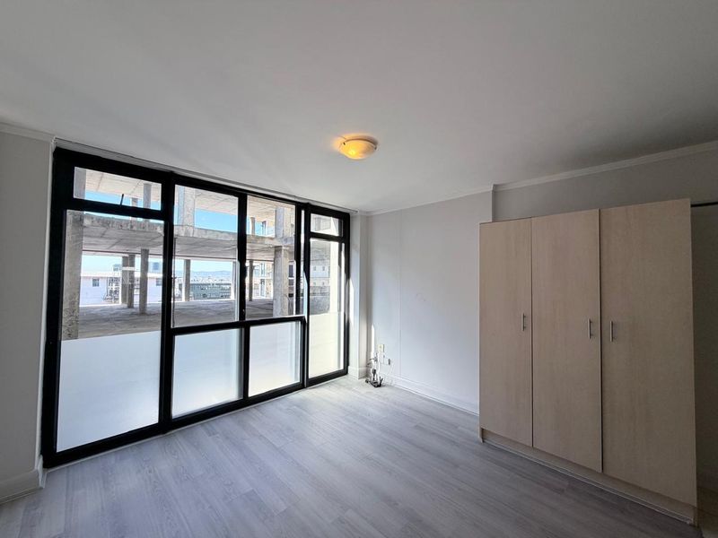 A bright bedroom featuring modern laminate flooring, a built-in wardrobe, and large windows offering an urban view.
