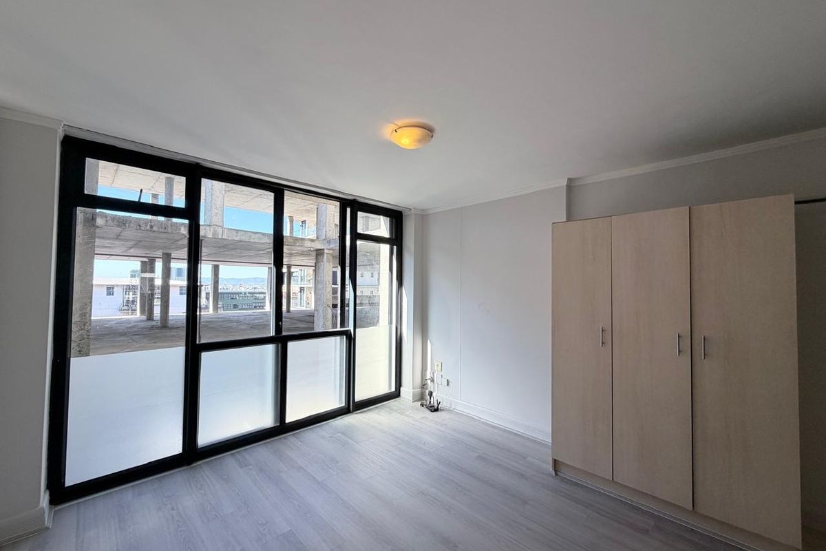A bright bedroom featuring modern laminate flooring, a built-in wardrobe, and large windows offering an urban view.