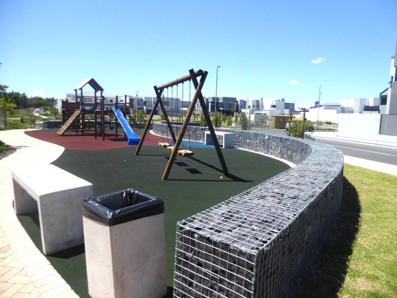 A well-maintained communal playground area featuring modern play equipment, a curved gabion retaining wall, and concrete seating within a residential estate.