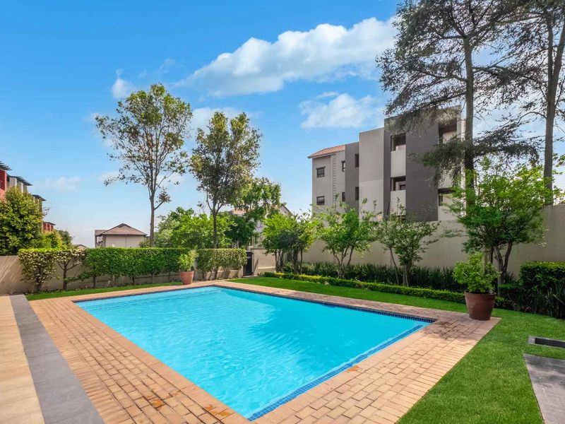 A well-maintained outdoor pool area featuring a clear swimming pool, paved surround, and landscaped garden borders within a secure apartment complex setting.