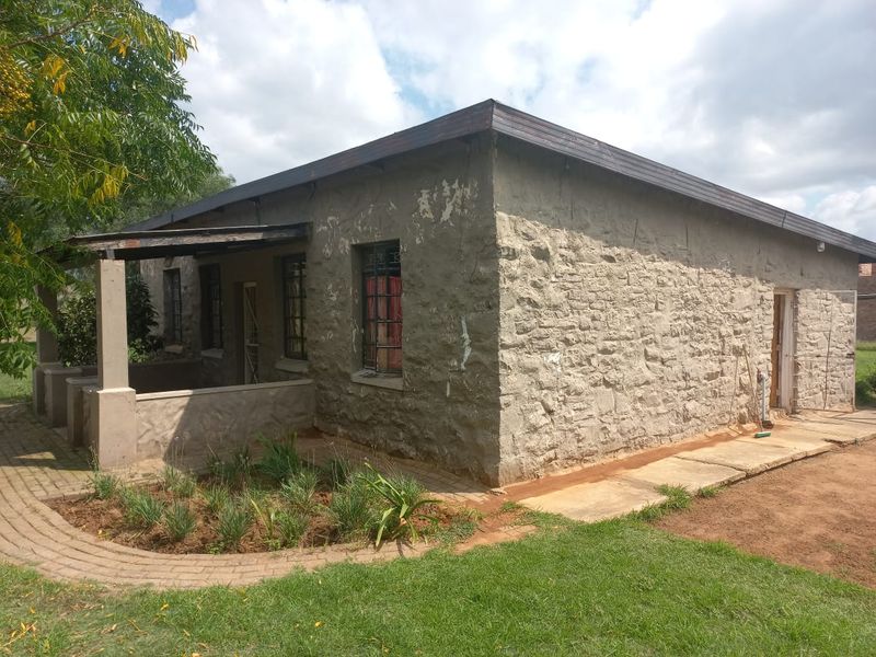 Rear view of a stone-walled house featuring a covered patio, paved walkway, and metal security bars on windows.
