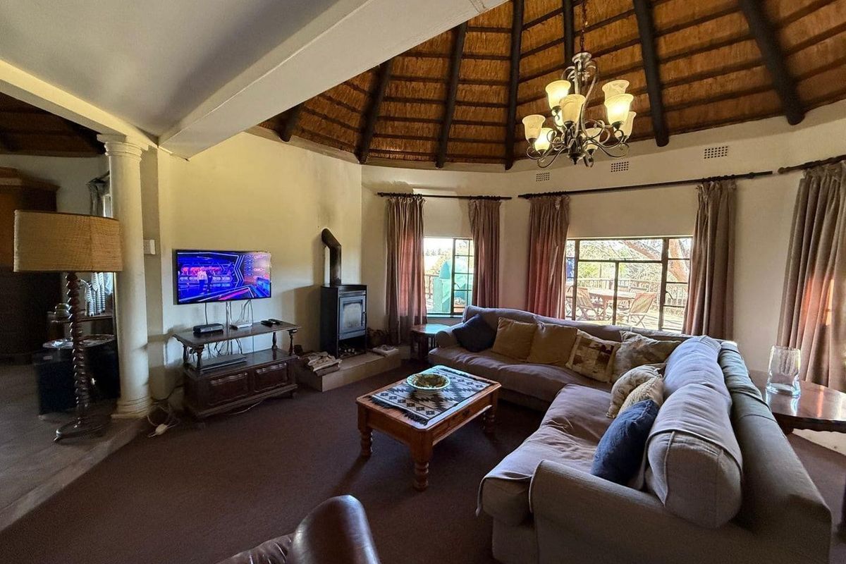 Spacious living room featuring a high thatched ceiling, exposed timber beams, a built-in fireplace, and large windows providing natural light and garden access.
