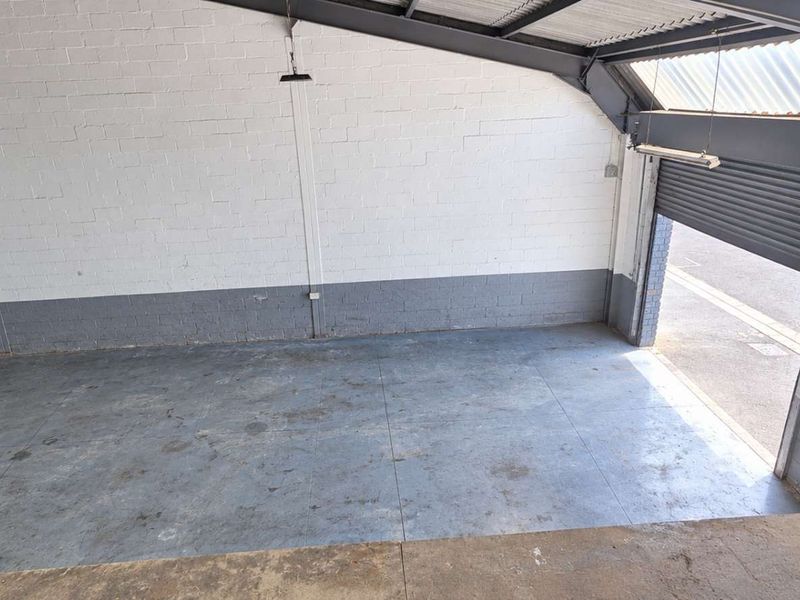 Spacious industrial warehouse or garage unit featuring high ceilings with exposed steel trusses, concrete flooring, and a large roller shutter door for vehicle access.