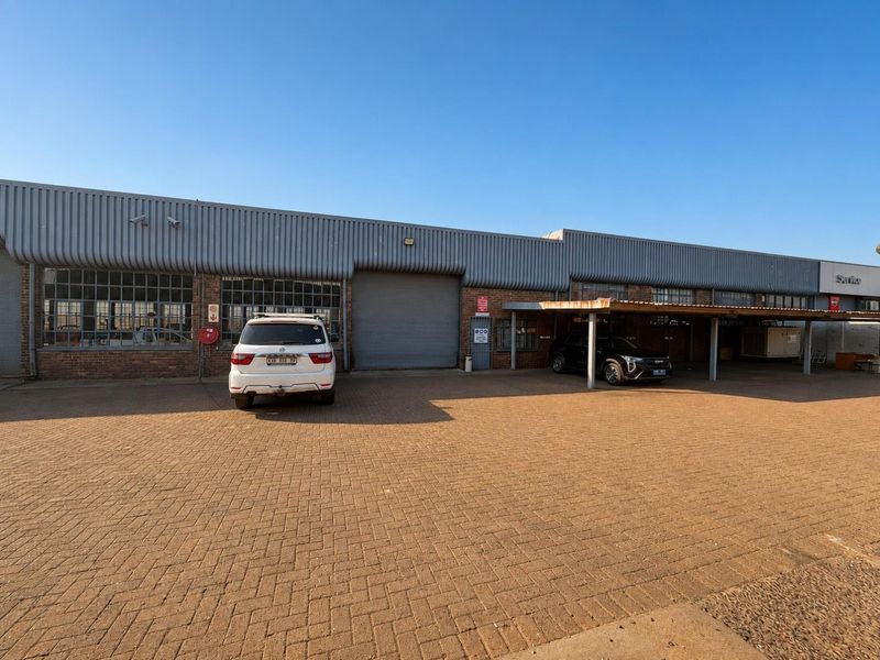 Front elevation of a commercial industrial property featuring a paved driveway, covered parking, roller shutter access, and brick and corrugated metal construction.