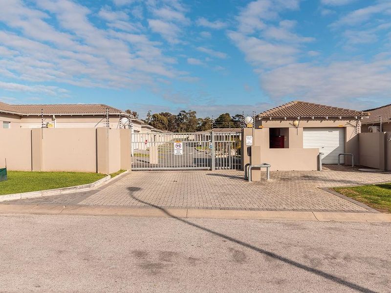 Front view of a secure townhouse complex entrance featuring a paved driveway, automated security gate, and perimeter wall with electric fencing.