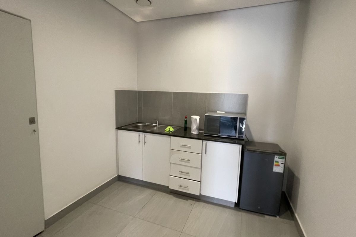 A compact kitchenette featuring modern white cabinetry, stone countertops, a stainless steel sink, and tiled finishes in a clean, neutral space.