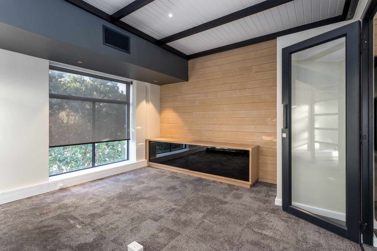 Modern office space featuring timber wall paneling, built-in cabinetry, exposed ceiling beams, and large windows with roller blinds.