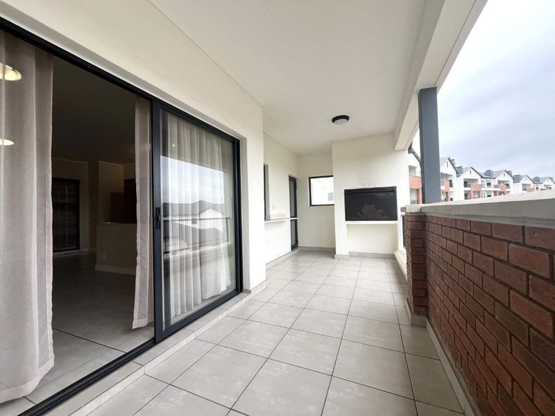 A spacious covered balcony featuring tiled flooring, a built-in braai, and a brick balustrade, offering seamless access to the interior living area.