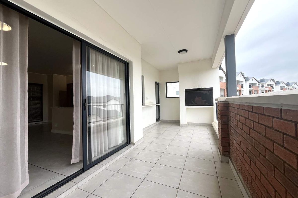 A spacious covered balcony featuring tiled flooring, a built-in braai, and a brick balustrade, offering seamless access to the interior living area.