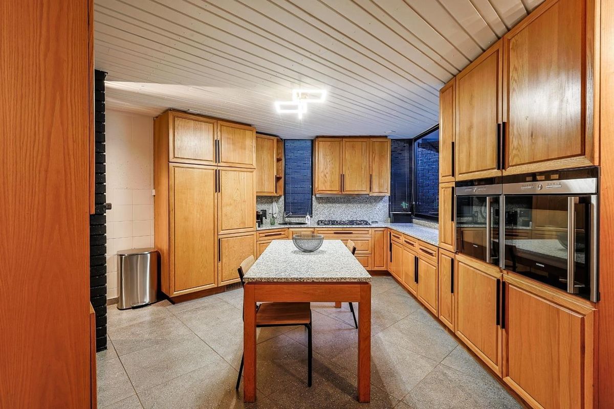 A functional kitchen featuring extensive wooden cabinetry, granite countertops, a central island, and built-in double ovens, set against a tiled floor and paneled ceiling.