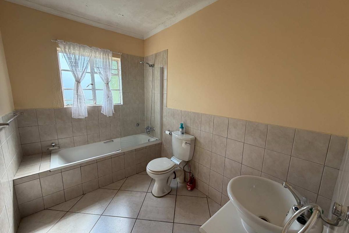 A functional bathroom featuring a built-in bathtub with shower mixer, ceramic toilet, and a pedestal basin with tiled flooring and partial wall tiling.