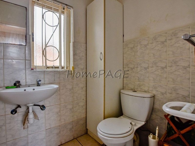 A dated bathroom featuring a pedestal basin, toilet, and built-in storage cupboard, with tiled walls and a security-gated window.