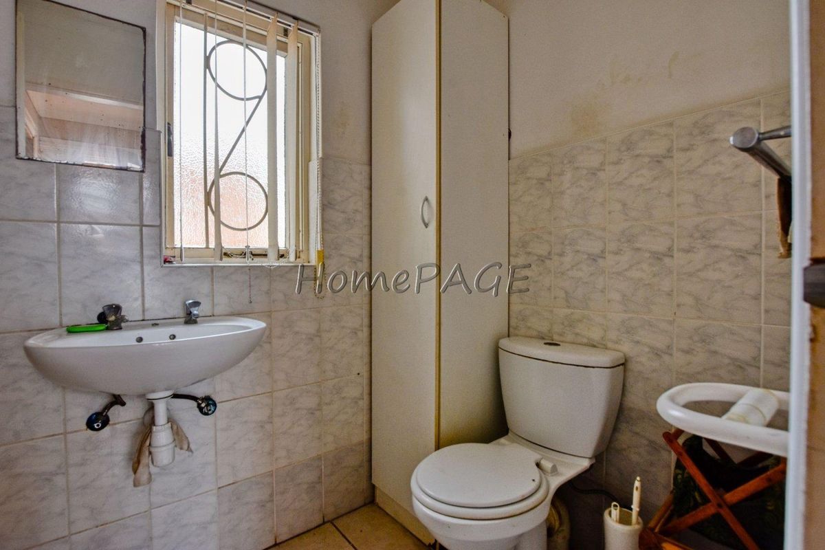 A dated bathroom featuring a pedestal basin, toilet, and built-in storage cupboard, with tiled walls and a security-gated window.