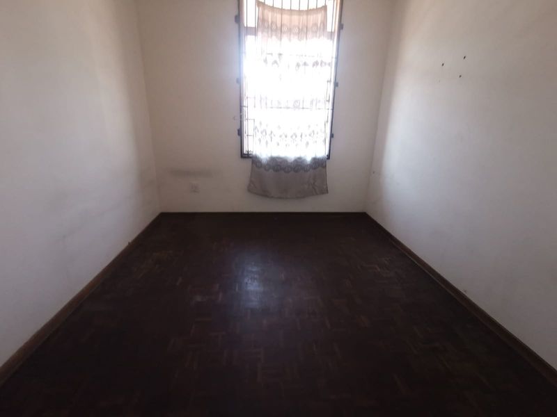 An empty bedroom featuring dark parquet flooring and a window fitted with security bars, requiring cosmetic updates to the wall surfaces.