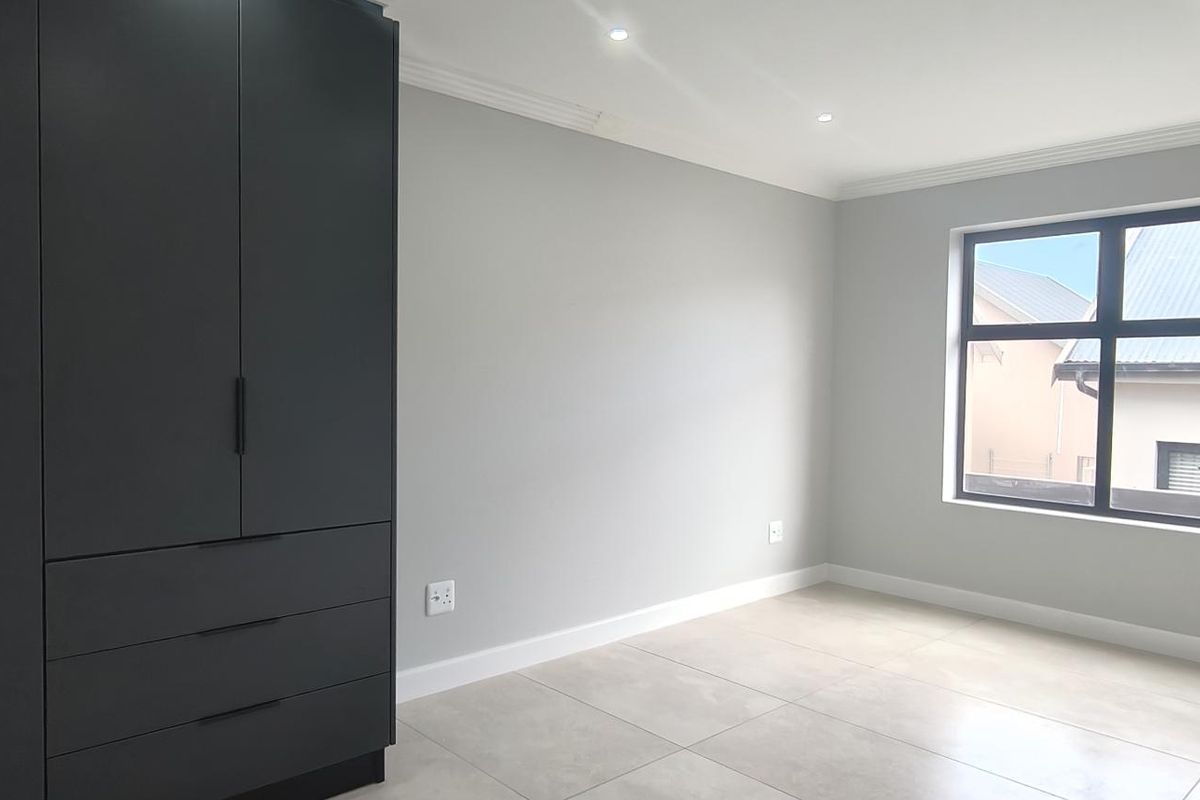 A bright, modern bedroom featuring neutral tiled flooring, recessed ceiling lighting, and a large, dark built-in wardrobe.