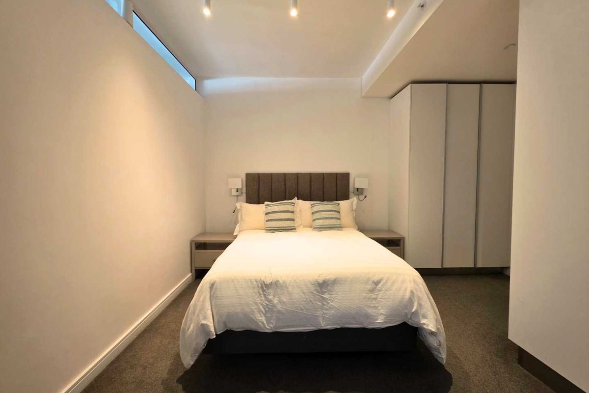 A modern bedroom featuring built-in wardrobes, track lighting, and a high-level clerestory window, providing a clean and functional living space.