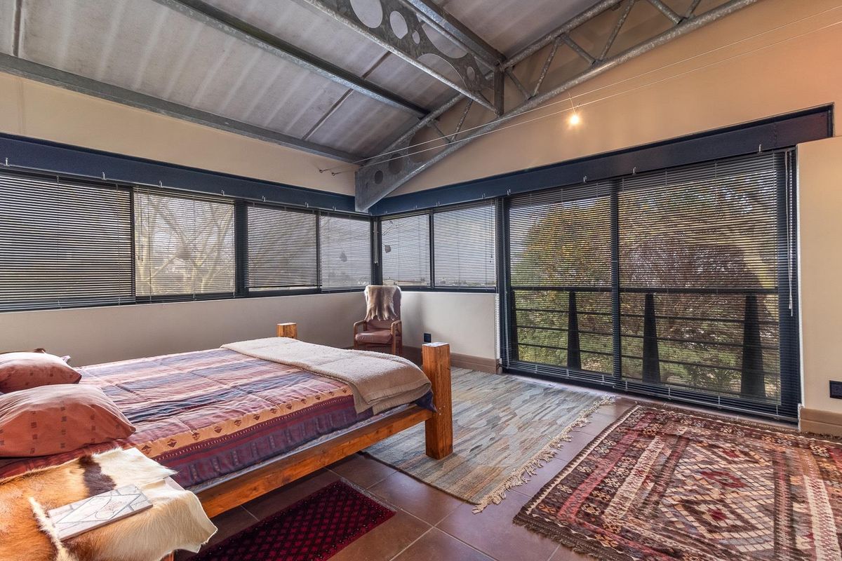 Spacious bedroom featuring industrial-style exposed steel roof trusses, tiled flooring, and large sliding glass doors with venetian blinds providing ample natural light.