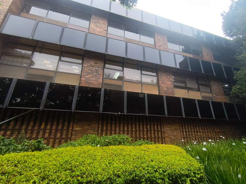 A multi-story office building facade featuring classic brickwork and extensive tinted glass windows, set behind a well-maintained landscaped garden.