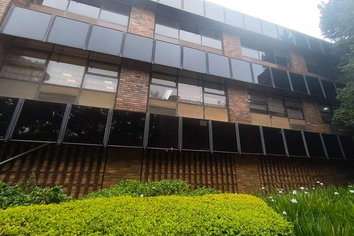 A multi-story office building facade featuring classic brickwork and extensive tinted glass windows, set behind a well-maintained landscaped garden.