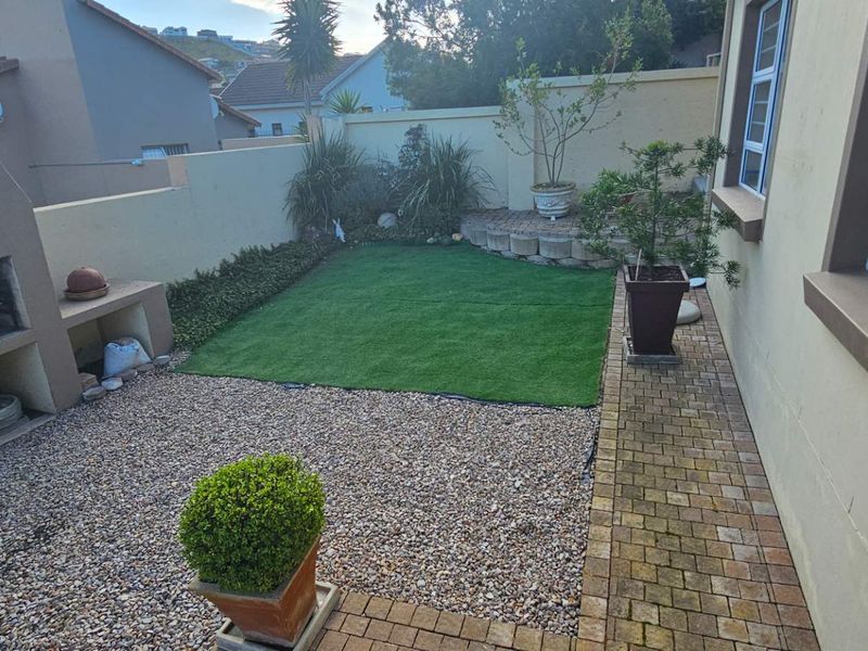 A low-maintenance rear garden featuring a built-in braai, artificial lawn, gravel area, and paved walkway enclosed by a boundary wall.