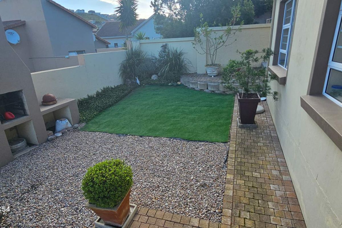 A low-maintenance rear garden featuring a built-in braai, artificial lawn, gravel area, and paved walkway enclosed by a boundary wall.