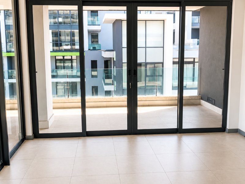 A bright, empty living area featuring large sliding glass doors that open onto a balcony, with modern tiled flooring and recessed ceiling lighting.