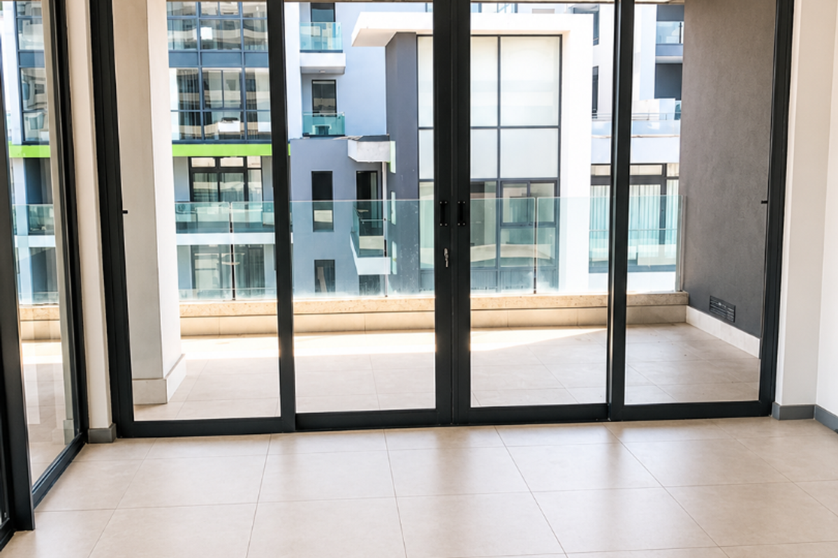A bright, empty living area featuring large sliding glass doors that open onto a balcony, with modern tiled flooring and recessed ceiling lighting.