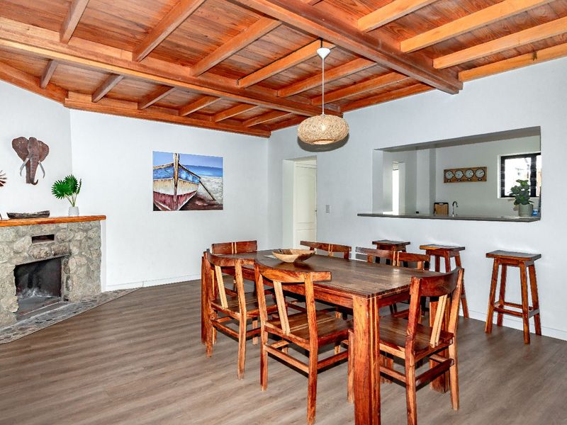 Open plan dining area featuring a rustic stone fireplace, exposed timber beam ceiling, and a breakfast bar overlooking the kitchen.