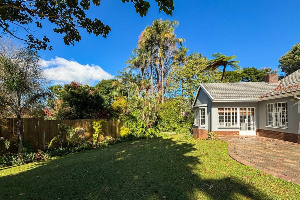A well-maintained rear garden featuring a paved patio area, lush lawn, mature trees, and a brick-accented exterior wall with classic window detailing.