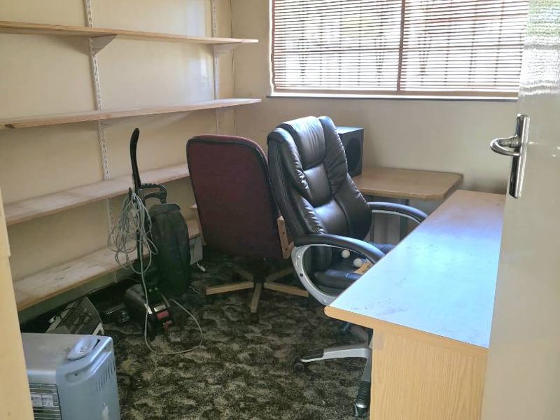 A small home office featuring built-in wall shelving, a window with blinds, and carpeted flooring, currently in need of cosmetic updates.