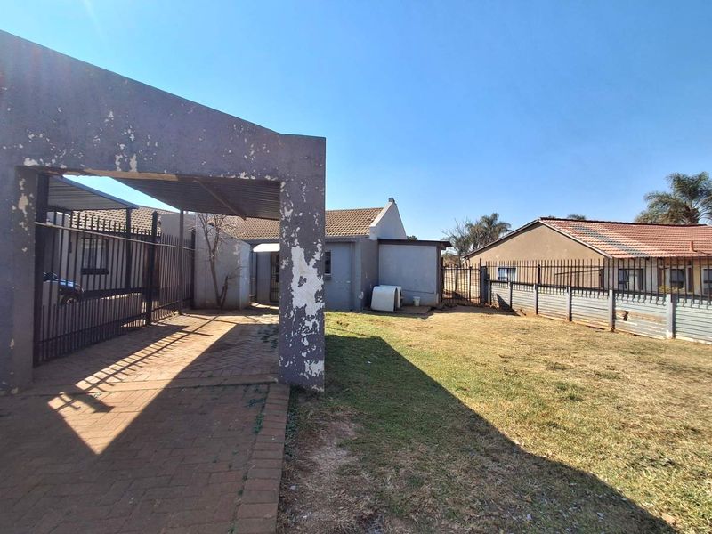 Rear exterior view of the property featuring a paved carport, metal perimeter fencing, and a spacious lawn area.