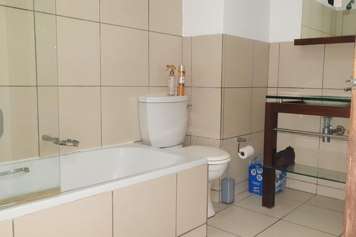 A functional bathroom featuring a bathtub with a glass screen, a toilet, and a vanity unit with glass shelving and neutral wall tiling.