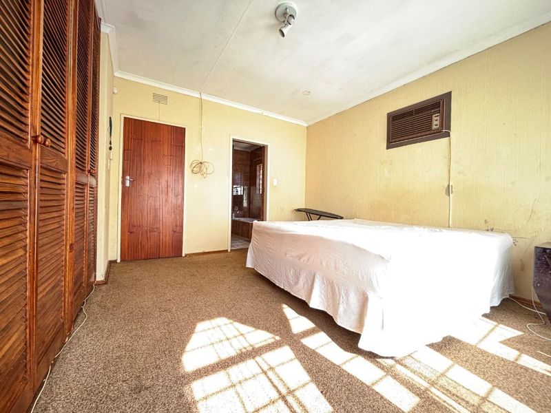 A bedroom featuring built-in wooden cupboards, carpeted flooring, and a wall-mounted air conditioning unit, with an adjoining bathroom visible through the doorway.