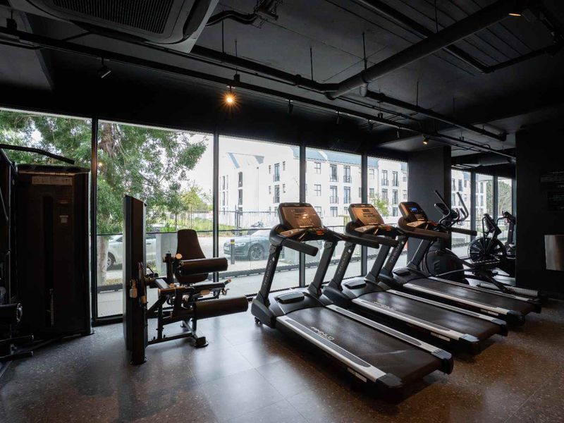 Modern apartment complex gym featuring floor-to-ceiling windows, exposed industrial-style ceiling ducting, and high-quality tiled flooring.