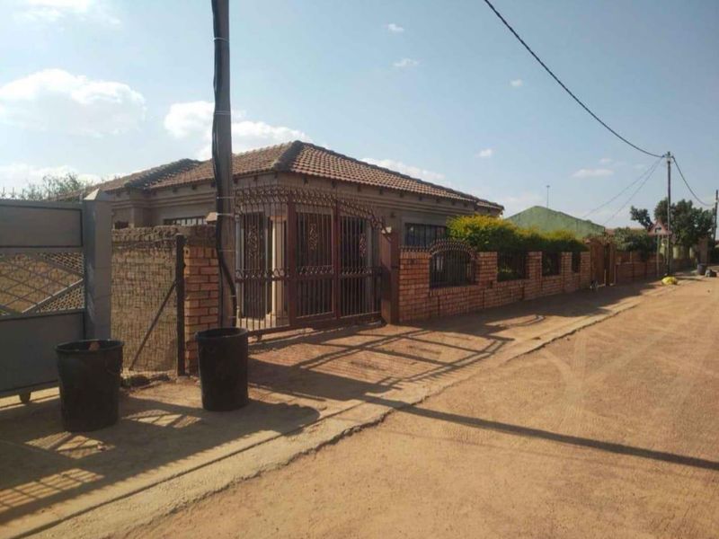 Front exterior view of a residential property featuring a brick boundary wall, wrought iron gate, and tiled roof, situated along an unpaved road.