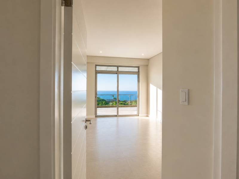 A bright, empty bedroom featuring neutral tiled flooring, recessed lighting, and a large sliding glass door leading to a balcony with ocean views.
