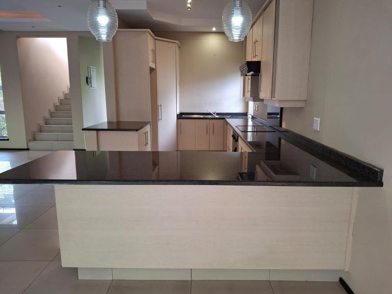 A spacious, empty kitchen featuring light-toned cabinetry, dark granite countertops, and tiled flooring, with an open-plan layout leading to a staircase.