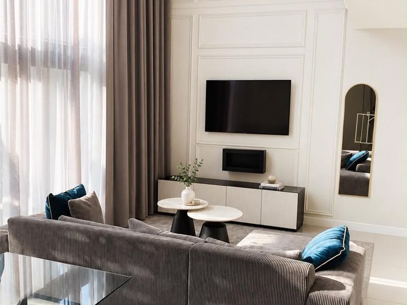 A modern, double-volume living room featuring elegant wall paneling, large floor-to-ceiling windows with floor-length curtains, and sleek tiled flooring.