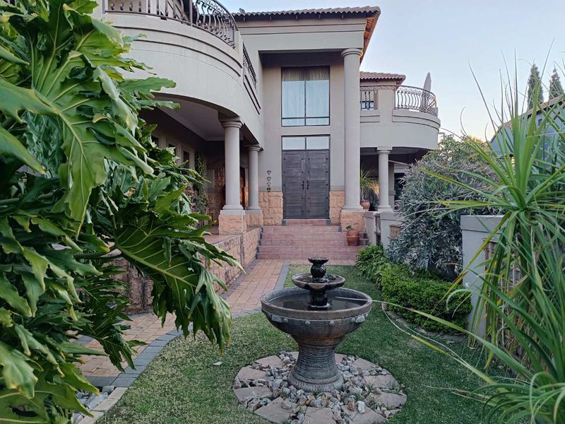 A grand double-story residence featuring a stone-clad base, classic columns, arched balconies, and a formal entrance with a landscaped garden and water feature.