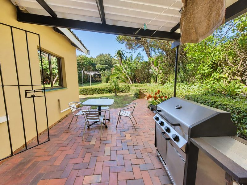 A covered outdoor patio featuring brick paving and a built-in braai area, overlooking a lush, established garden.