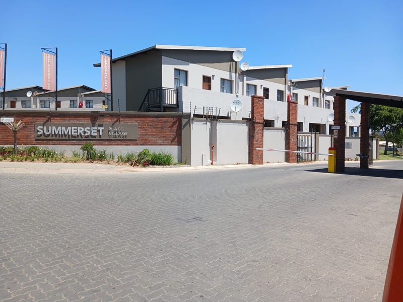 Front entrance view of the Summerset Village townhouse complex featuring a paved driveway, security boom gate, and brick boundary wall with electric fencing.