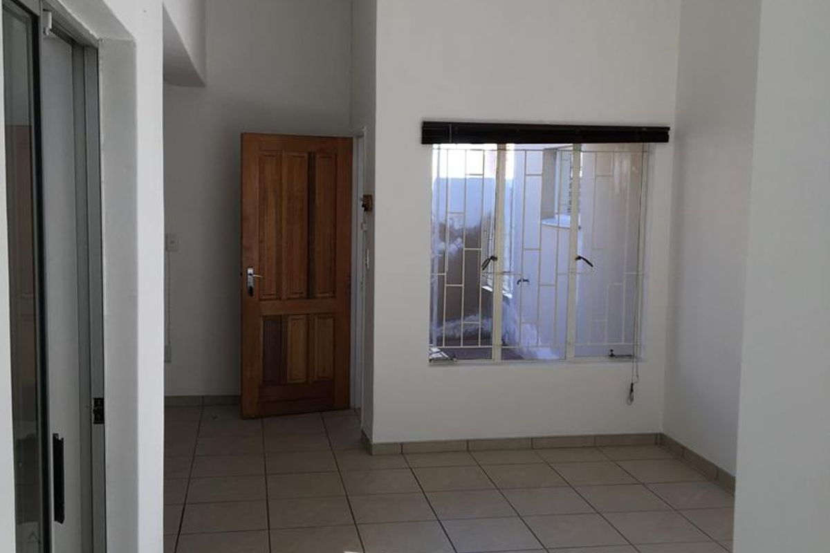 A bright, empty living area featuring neutral floor tiles, a wooden door, and a window fitted with security bars.