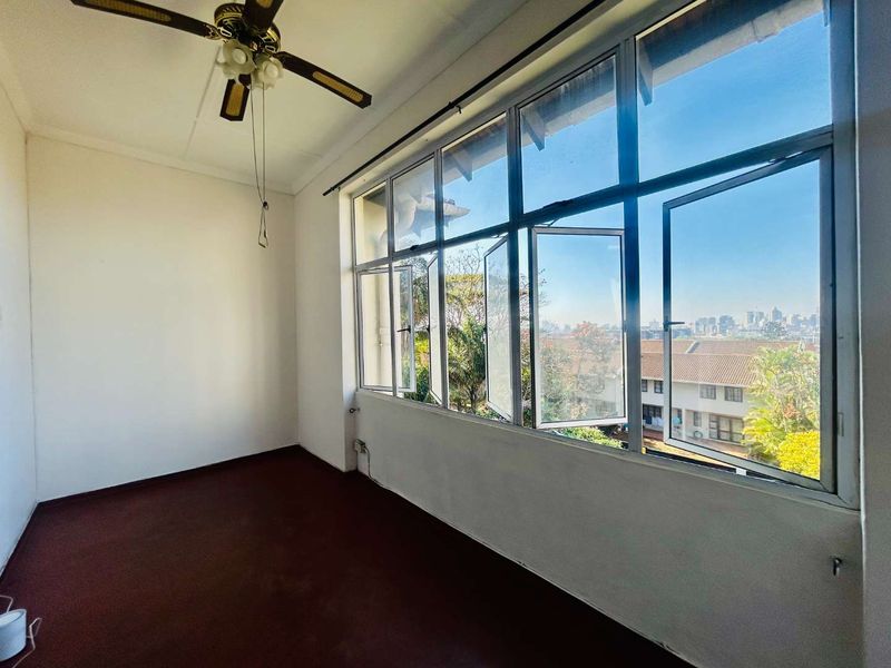 An enclosed balcony or sunroom featuring large aluminium-framed windows with expansive views, a ceiling fan, and carpeted flooring.