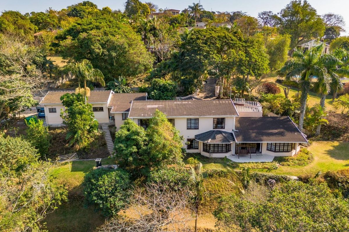 Aerial view of a residential property featuring a multi-section tiled roof, established garden, and a terrace area set within a lush, green landscape.