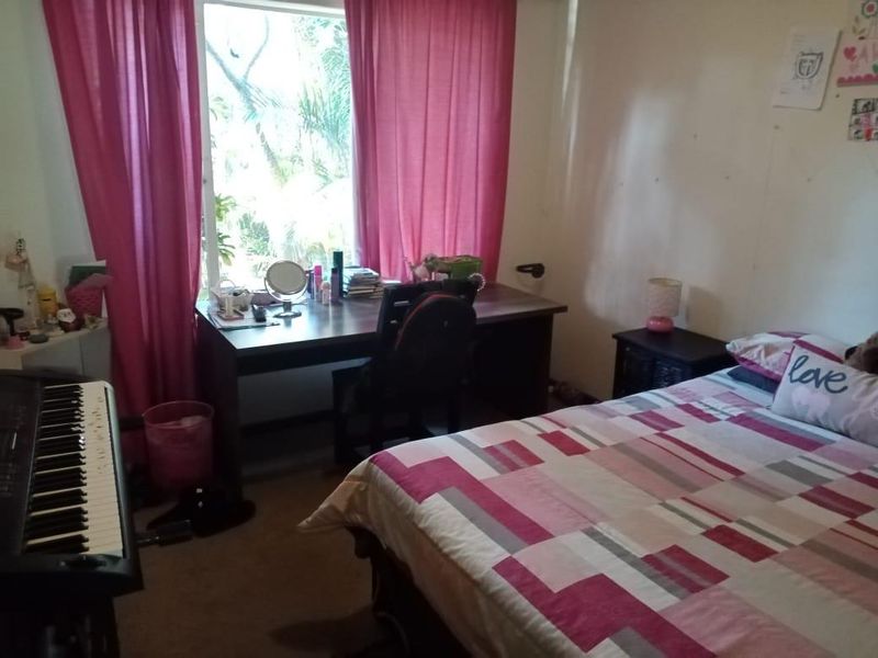A bedroom featuring carpet flooring and a large window providing natural light, currently used as a combined sleeping and study area.