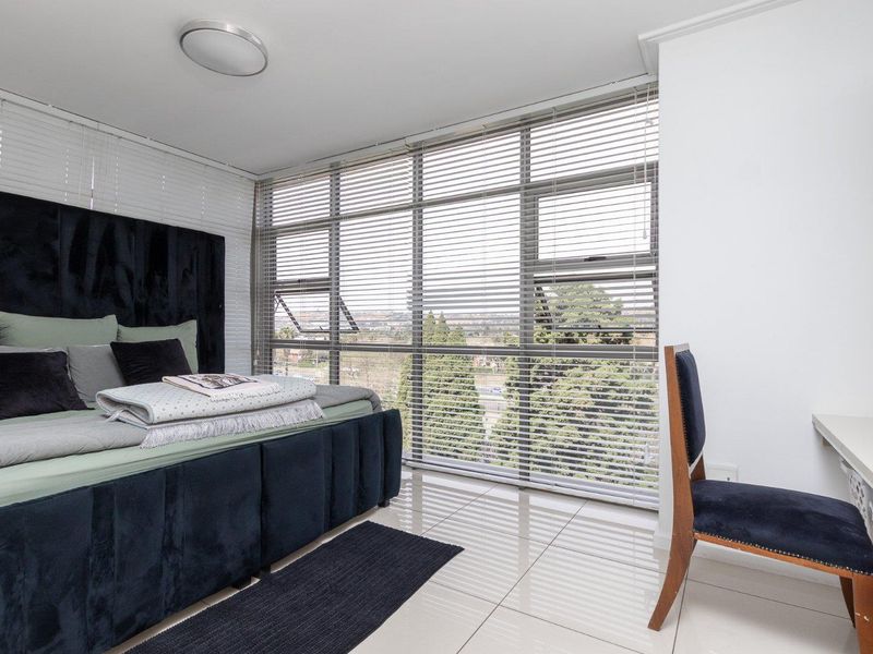 A bright bedroom featuring large floor-to-ceiling windows with venetian blinds, tiled flooring, and a built-in desk area.