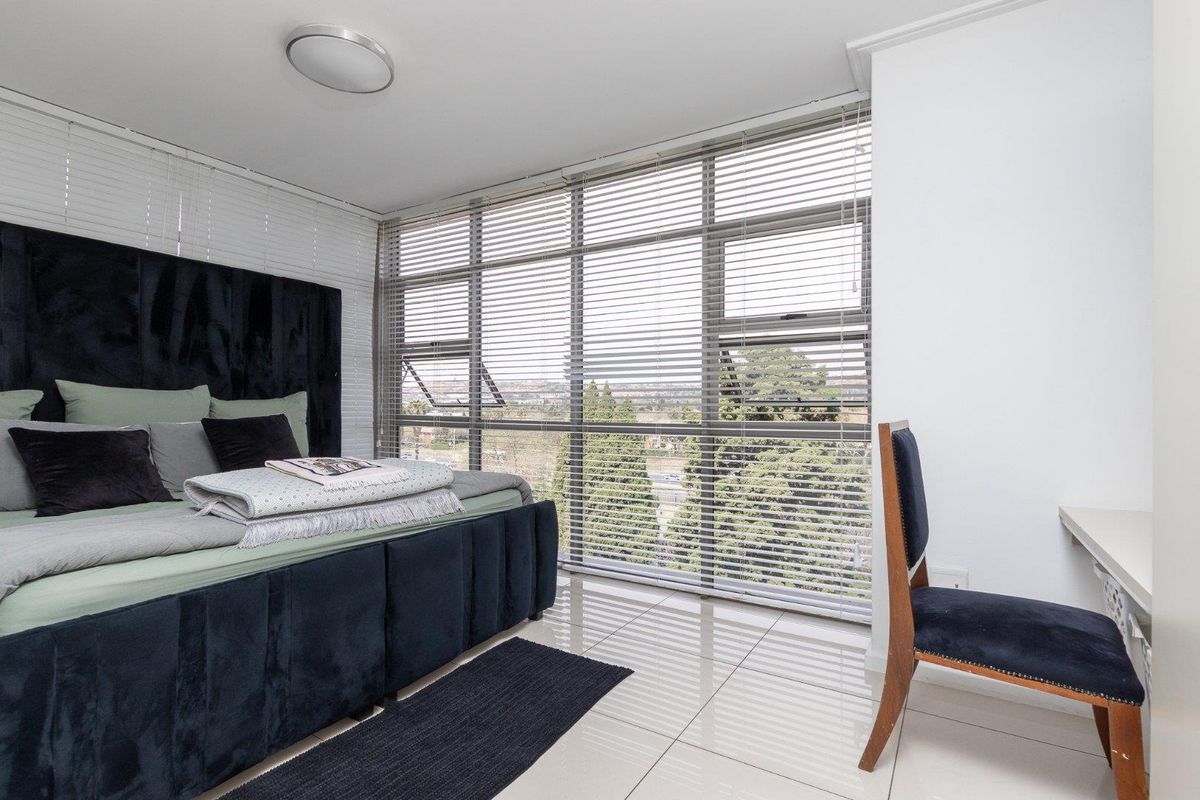 A bright bedroom featuring large floor-to-ceiling windows with venetian blinds, tiled flooring, and a built-in desk area.
