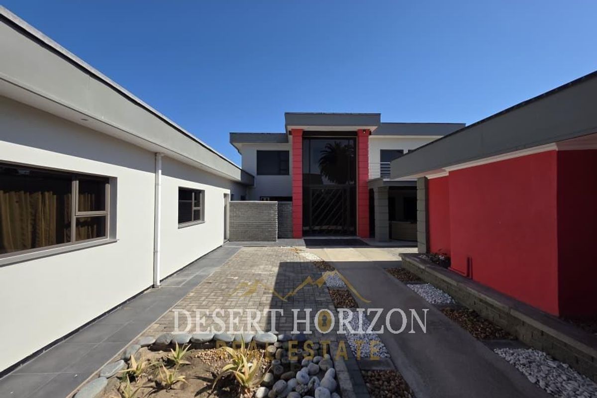 A modern rear courtyard featuring paved surfaces, landscaped garden beds, and a striking red-accented entrance facade with large glass doors.