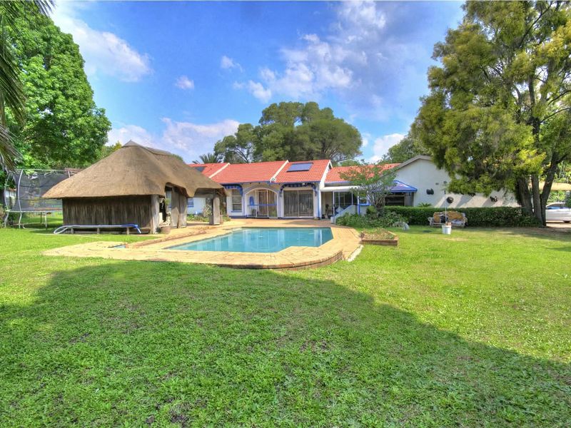 Rear view of the property featuring a swimming pool, a traditional thatch lapa, and a spacious garden area with mature trees.